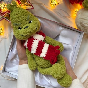 May include: A green Grinch doll with a red and white striped scarf, presented in a white gift box. The doll has a smiling face and is made of a soft, textured material. The background features Christmas lights.