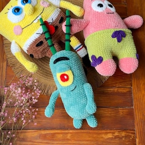 May include: Three plush toys made of soft yarn featuring a yellow rectangular character with blue eyes, a pink star-shaped character wearing green shorts with purple flowers, and a small teal character with one red eye and two long antennae.