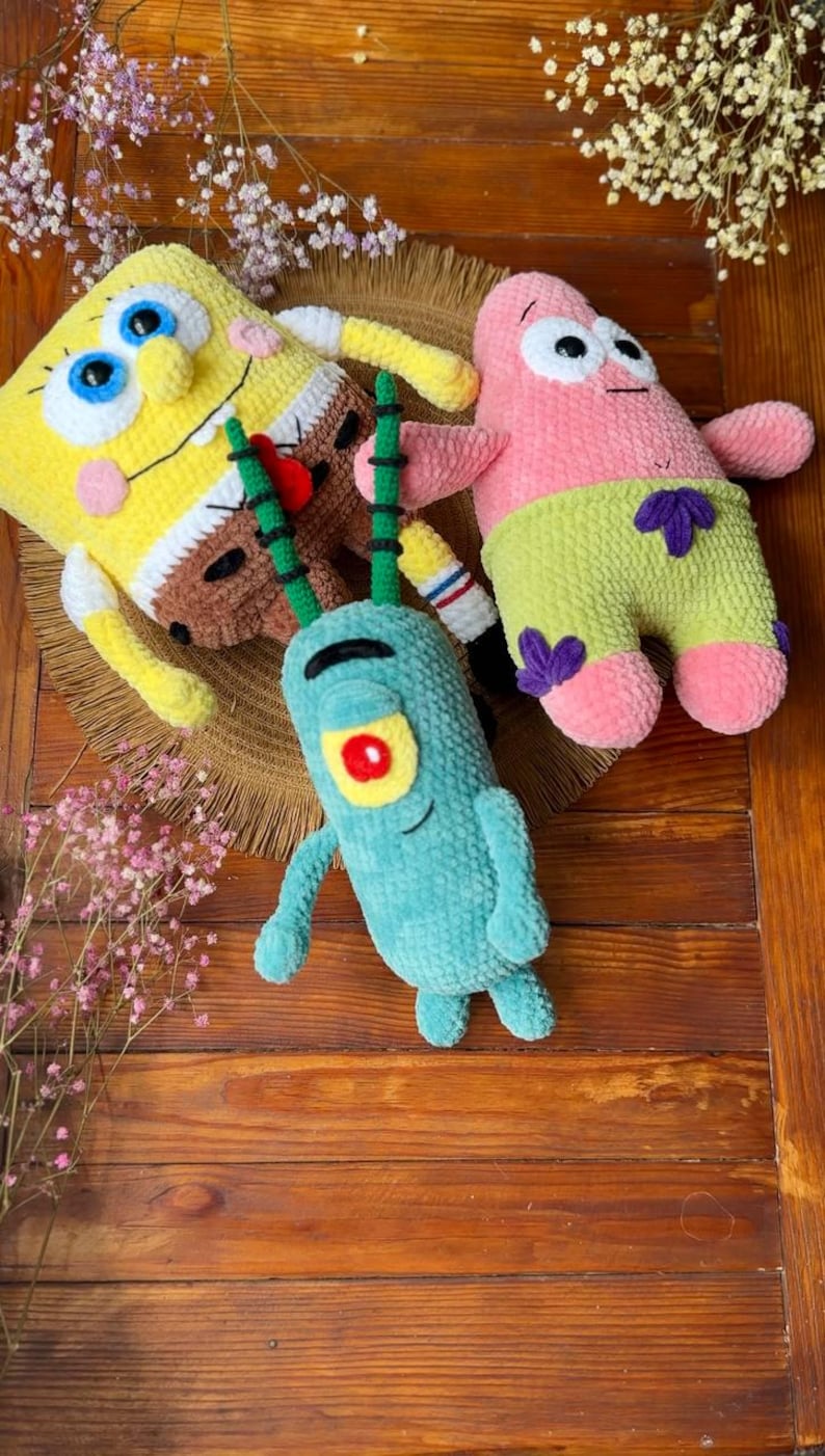 May include: Three plush toys made of soft yarn featuring a yellow rectangular character with blue eyes, a pink star-shaped character wearing green shorts with purple flowers, and a small teal character with one red eye and two long antennae.