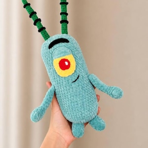 May include: A teal crochet plush toy shaped like a one-eyed character with two long antennae, a single yellow eye with a red pupil, and a small black embroidered smile. The textured yarn body features two thin arms and two short legs.