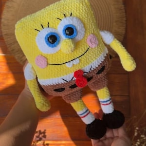 May include: A yellow rectangular crochet plush toy resembling a cartoon character with large blue eyes, a wide smile, pink cheeks, and a small nose. The figure wears brown shorts, white socks with red and blue stripes, and black shoes.