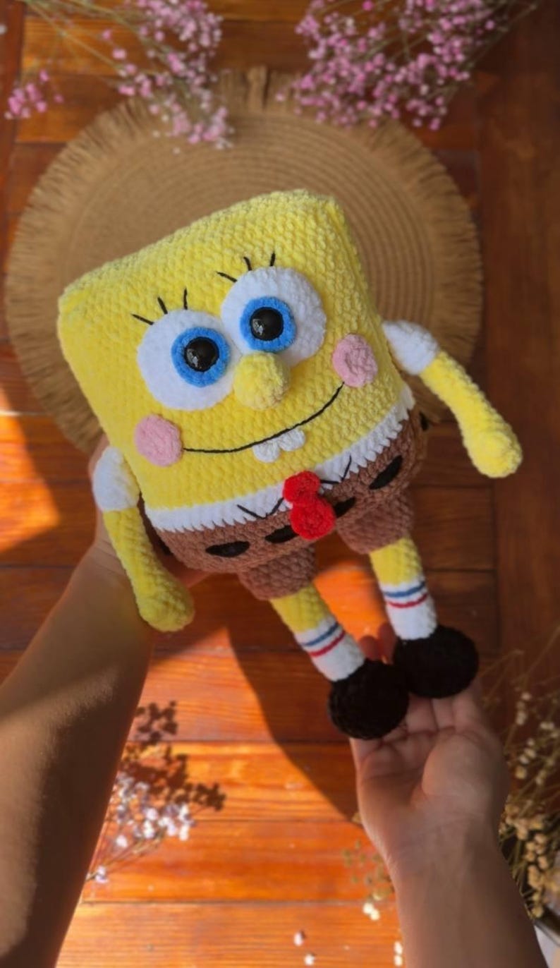 May include: A yellow rectangular crochet plush toy resembling a cartoon character with large blue eyes, a wide smile, pink cheeks, and a small nose. The figure wears brown shorts, white socks with red and blue stripes, and black shoes.