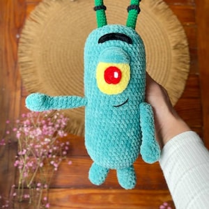May include: A teal crochet plush toy shaped like a single-eyed character with two long green antennae featuring black stripes. The figure has a yellow eye patch with a red center, a black embroidered eyebrow, and a small black stitched smile.