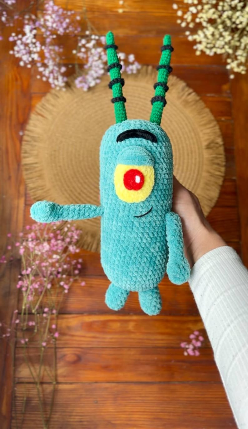May include: A teal crochet plush toy shaped like a single-eyed character with two long green antennae featuring black stripes. The figure has a yellow eye patch with a red center, a black embroidered eyebrow, and a small black stitched smile.
