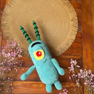 May include: A teal crochet plush toy shaped like a small, cylindrical creature with two long antennae, a single large yellow eye with a red pupil, and a thin black embroidered mouth. The plush has two small arms and two short legs.