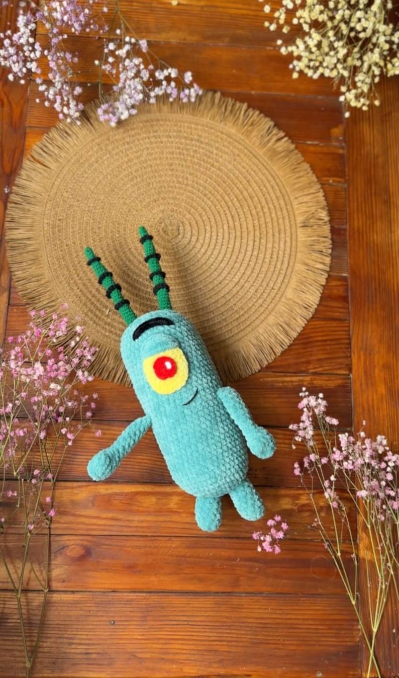 May include: A teal crochet plush toy shaped like a small, cylindrical creature with two long antennae, a single large yellow eye with a red pupil, and a thin black embroidered mouth. The plush has two small arms and two short legs.