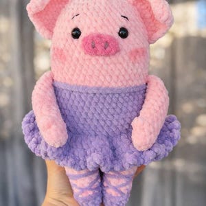May include: A handmade pink and purple crochet piglet wearing a purple tutu and pink ballet slippers. The piglet has pink ears, a snout, and rosy cheeks. The plush toy is soft and cuddly.