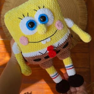 May include: A yellow plush toy shaped like a rectangular character with large blue eyes, a wide smile, and two white teeth. It wears brown shorts, white socks with red and blue stripes, and black shoes. The toy is crafted from soft yarn.