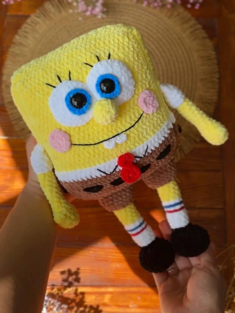 May include: A yellow plush toy shaped like a rectangular character with large blue eyes, a wide smile, and two white teeth. It wears brown shorts, white socks with red and blue stripes, and black shoes. The toy is crafted from soft yarn.