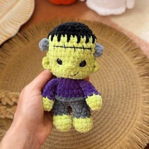May include: A handmade crochet Frankenstein's monster doll with a green face, black hair, and a purple shirt. The doll has gray pants and small gray ears. Other crochet Halloween figures are in the background.