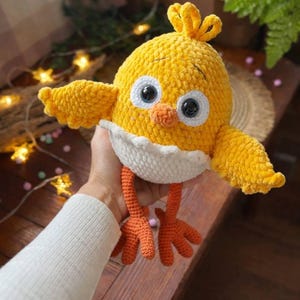 May include: A handmade, yellow crochet chick with large, black and white eyes, orange beak and feet, and small wings. The chick has a white belly and is a soft, plush toy. The chick is a cute, whimsical design.