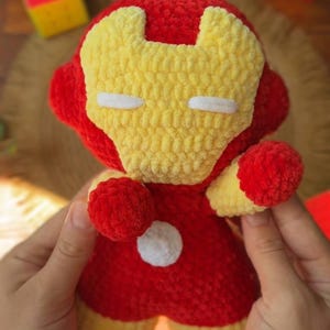 May include: A plush toy crafted from red and yellow yarn, designed to resemble a superhero character. The figure features a yellow face mask, red helmet, and a white circular accent on the chest. The texture is soft and fuzzy, with rounded limbs.