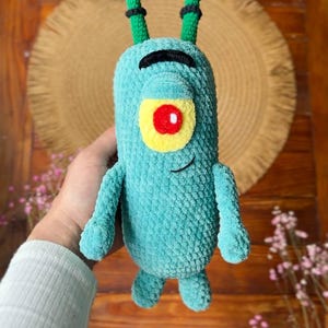 May include: A teal crochet plush toy featuring a single large eye with a yellow sclera and red pupil, a black arched eyebrow, and a small stitched smile. The character has two long, thin, green antennae with black horizontal stripes and two small arms.