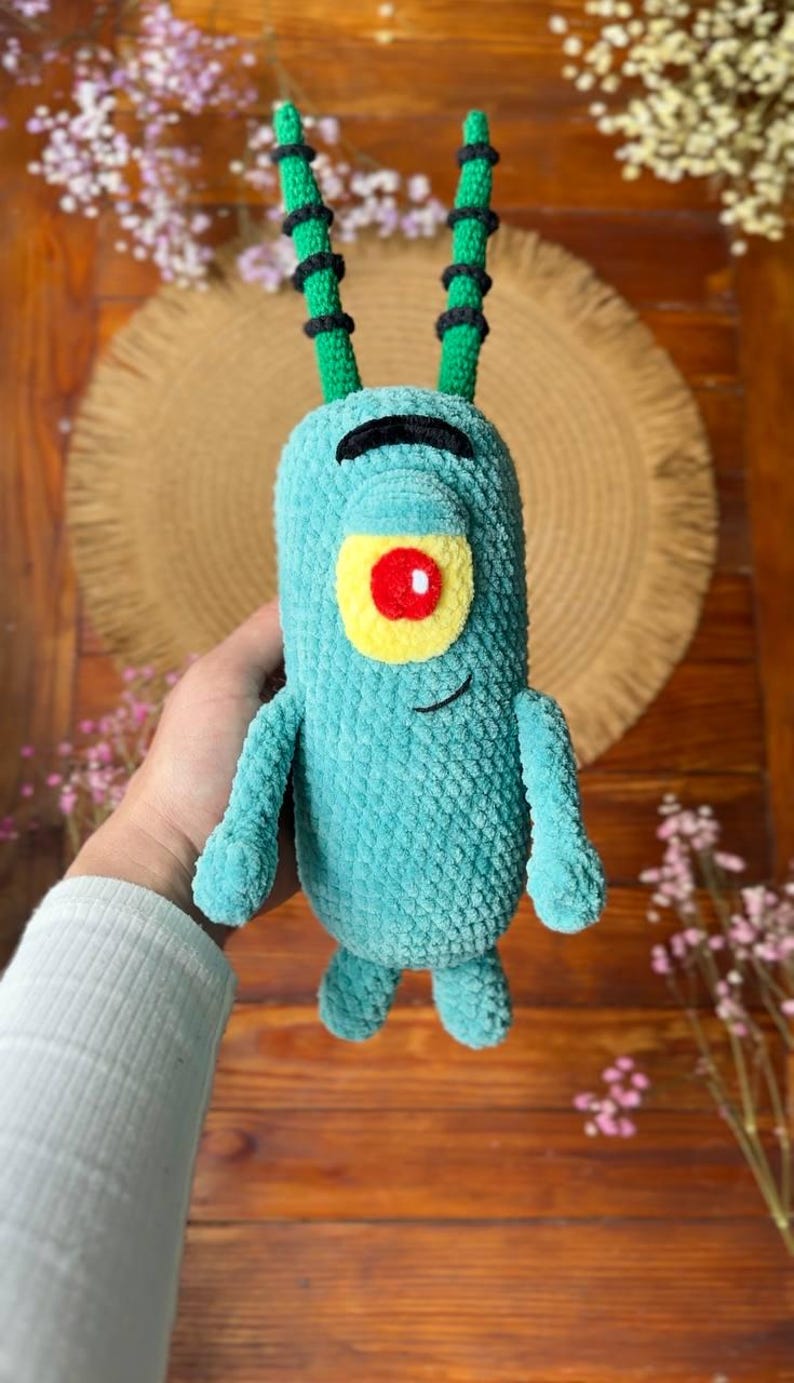 May include: A teal crochet plush toy featuring a single large eye with a yellow sclera and red pupil, a black arched eyebrow, and a small stitched smile. The character has two long, thin, green antennae with black horizontal stripes and two small arms.