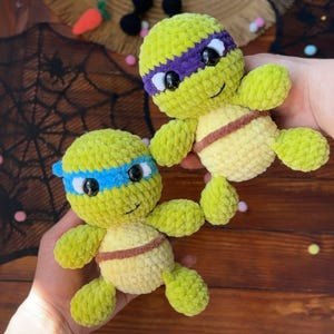 May include: Two handmade, plush turtle toys in the style of Teenage Mutant Ninja Turtles. The turtles are green with yellow bellies, brown belts, and different colored headbands. One has a blue headband, the other a purple one.