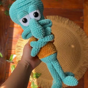 May include: A handmade crochet plush toy of a teal-colored character with a large head, prominent nose, and white eyes with black pupils. The figure wears a mustard-yellow shirt and has long, thin arms and legs crafted from soft, textured yarn.