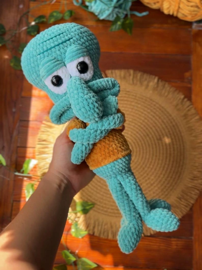May include: A handmade crochet plush toy of a teal-colored character with a large head, prominent nose, and white eyes with black pupils. The figure wears a mustard-yellow shirt and has long, thin arms and legs crafted from soft, textured yarn.