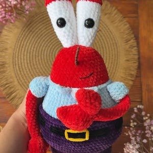 Op de afbeelding: Een gehaakt knuffeltje van een rood krabpersonage met hoge witte oogstelen, zwarte knoopogen, een lichtblauw overhemd en een donkerpaarse broek met een gele riemgesp. Het figuurtje heeft rode scharen en een kleine bruine geborduurde mond.