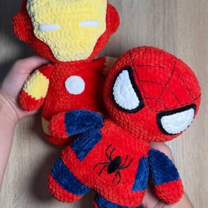 Puede incluir: Dos peluches hechos de hilo rojo, amarillo y azul. Uno presenta una máscara roja y amarilla con ojos blancos, representando un personaje robótico. El otro es rojo y azul con un emblema de araña negra en el pecho y ojos blancos.