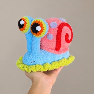 Puede incluir: Un juguete de caracol tejido a ganchillo con un cuerpo azul claro, una base de color verde lima texturizada y una concha rosa con manchas moradas y un patrón de espiral rojo. El caracol tiene dos ojos grandes y prominentes con detalles circulares naranjas, negros y blancos.