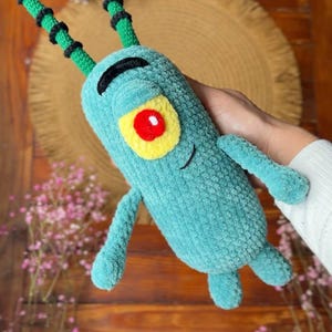 May include: A plush toy shaped like a small, teal, cylindrical creature with a single large red eye surrounded by a yellow patch. It features two long, green, segmented antennae with black bands, a black embroidered eyebrow, and a small smile.