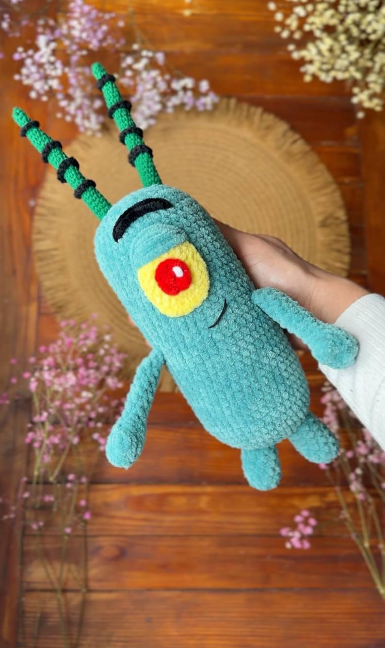 May include: A plush toy shaped like a small, teal, cylindrical creature with a single large red eye surrounded by a yellow patch. It features two long, green, segmented antennae with black bands, a black embroidered eyebrow, and a small smile.