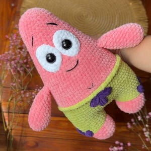 Puede incluir: Un peluche con forma de estrella de mar rosa de textura suave. Presenta dos grandes ojos circulares blancos con pupilas negras, una pequeña sonrisa bordada y pantalones cortos de color verde claro con detalles de flores moradas en las piernas.
