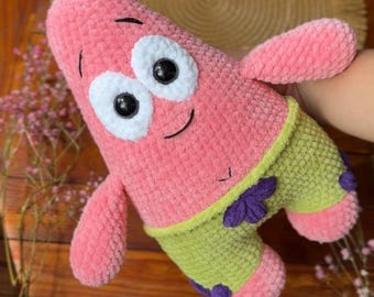 May include: A plush toy shaped like a pink starfish with a textured surface. It features two large white circular eyes with black pupils, a small stitched smile, and light green shorts with purple flower accents on the legs and waist.