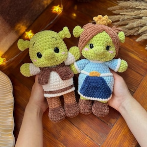 Può includere: Due bambole all'uncinetto fatte a mano, una che assomiglia a Shrek e l'altra a Fiona, vengono tenute in mano. La bambola di Shrek indossa un gilet marrone e pantaloni marroni. La bambola di Fiona indossa un vestito blu e una corona. Entrambe le bambole hanno la pelle verde e sorridono.