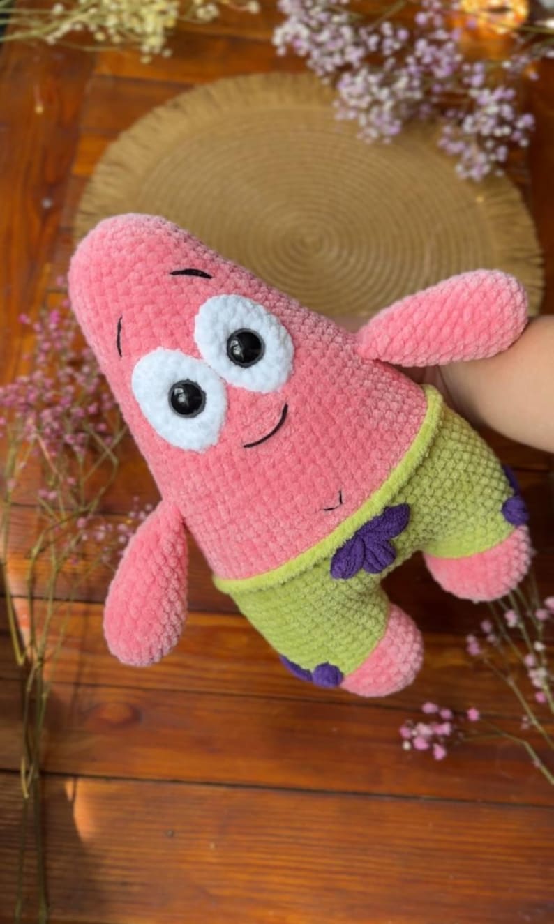 May include: A plush toy shaped like a pink starfish with a textured surface. It features large white circular eyes with black pupils, a stitched smile, and a small navel. The toy wears lime green shorts with purple flower accents on the waist.