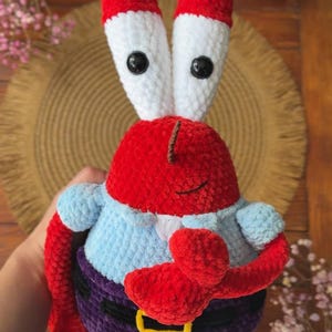 May include: A handmade crochet plush toy of a red crab character with tall white eye stalks, black button eyes, and brown antennae. The figure wears a light blue shirt, dark purple pants with a black belt, and a yellow buckle, featuring red pincers.
