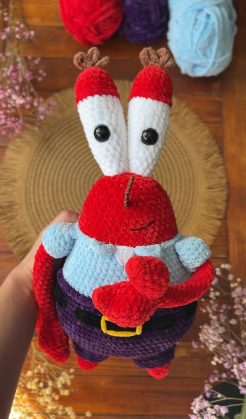May include: A handmade crochet plush toy of a red crab character with tall white eye stalks, black button eyes, and brown antennae. The figure wears a light blue shirt, dark purple pants with a black belt, and a yellow buckle, featuring red pincers.