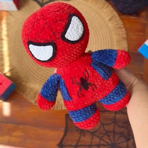 May include: A plush toy character made of red and blue yarn with a textured, crocheted finish. The figure features a round head with large white eyes outlined in black, and a black spider emblem embroidered on the center of the red chest.