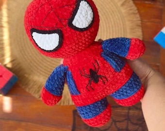 May include: A plush toy character made of red and blue yarn with a textured, crocheted finish. The figure features a round head with large white eyes outlined in black, and a black spider emblem embroidered on the center of the red chest.