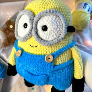 Puede incluir: Un juguete Minion tejido a mano. El Minion es amarillo con un peto azul, gafas grises y detalles negros. El juguete tiene dos ojos grandes y botones de madera en el peto. El fondo incluye hilo y un pequeño oso de peluche.