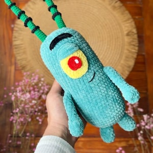 May include: A teal crochet plush toy shaped like a cartoon character with a single large yellow eye, a red pupil, two long green antennae with black rings, a black curved eyebrow, and a small stitched smile on its cylindrical body.