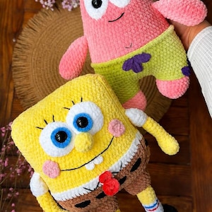May include: Two plush toys made of soft yarn. One is a pink, star-shaped character with green shorts featuring purple flower patterns. The other is a yellow, rectangular character with blue eyes, a white shirt, brown pants, and black shoes.