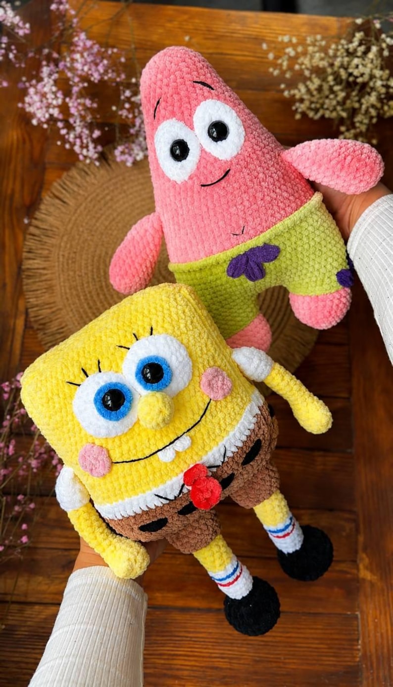 May include: Two plush toys made of soft yarn. One is a pink, star-shaped character with green shorts featuring purple flower patterns. The other is a yellow, rectangular character with blue eyes, a white shirt, brown pants, and black shoes.