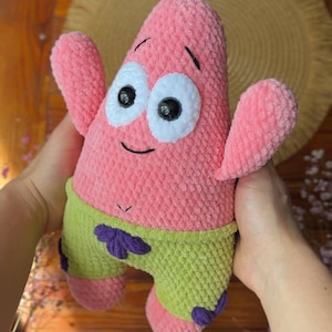 May include: A pink plush toy shaped like a starfish with large white eyes, black pupils, and a small smile. The character wears lime green shorts with purple flower accents. The texture of the plush is soft and fuzzy, featuring a rounded body.