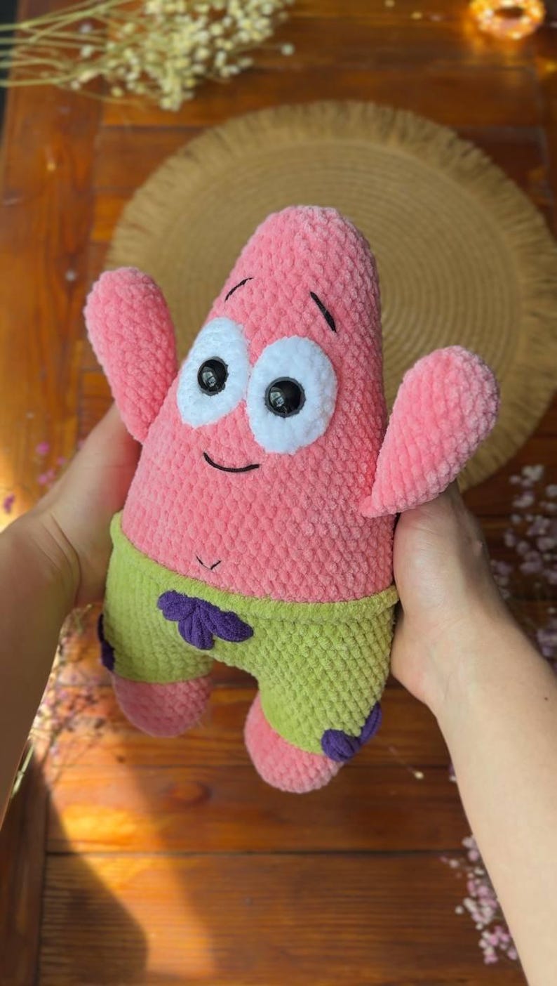 May include: A pink plush toy shaped like a starfish with large white eyes, black pupils, and a small smile. The character wears lime green shorts with purple flower accents. The texture of the plush is soft and fuzzy, featuring a rounded body.