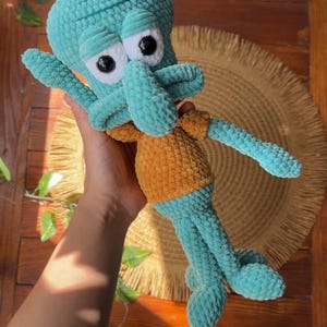 May include: A plush crochet toy of a teal-colored character with a large head, prominent nose, and wide forehead, wearing a mustard-yellow shirt. The figure features black plastic eyes set in white circular patches and has long, thin limbs.