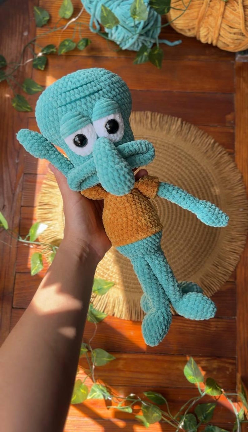 May include: A plush crochet toy of a teal-colored character with a large head, prominent nose, and wide forehead, wearing a mustard-yellow shirt. The figure features black plastic eyes set in white circular patches and has long, thin limbs.