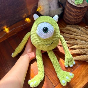 May include: A handmade, lime green crochet monster with one large eye, small horns, and long arms and legs. The monster has a white eye with a dark green center and a small smile. The feet have white accents. The toy is on a wooden surface.