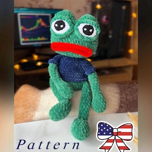 Può includere: Una bambola a forma di rana all'uncinetto verde fatta a mano con una camicia blu e labbra rosse. La bambola ha grandi occhi bianchi con pupille nere. La parola "Pattern" è scritta in blu. È visibile anche un fiocco con la bandiera americana.