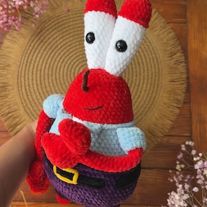 May include: A plush crochet toy of a red crab character with tall white eye stalks, black button eyes, and small brown antennae. The figure wears a light blue shirt, dark purple pants, and a black belt with a yellow buckle, crafted from yarn.
