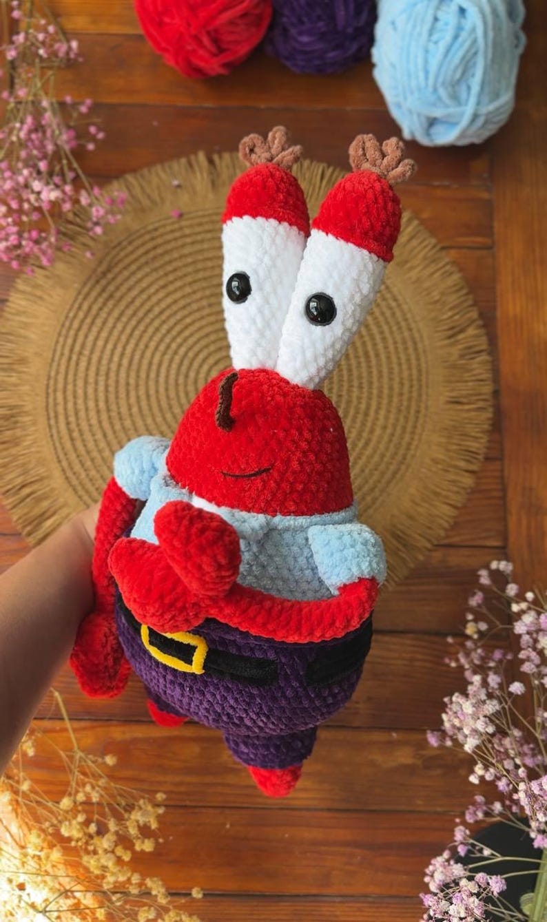 May include: A plush crochet toy of a red crab character with tall white eye stalks, black button eyes, and small brown antennae. The figure wears a light blue shirt, dark purple pants, and a black belt with a yellow buckle, crafted from yarn.