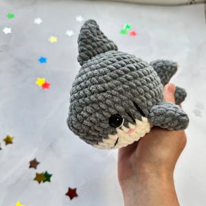 May include: A handmade grey crochet shark toy with black safety eyes and a white belly. The shark has a small pink mouth and is held in a hand. The background is white with colourful star-shaped confetti.