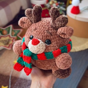 May include: A handmade brown plush reindeer with a red nose, black eyes, and brown antlers. It wears a red and green striped scarf. The reindeer is a soft, cuddly toy, perfect for holiday decor or as a gift.