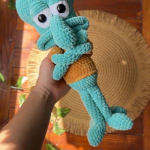 May include: A handmade crochet plush toy of a teal-colored character with a large head, prominent nose, and crossed arms. The figure features black and white plastic eyes and wears a mustard yellow shirt. The texture is soft, fuzzy, and yarn-based.
