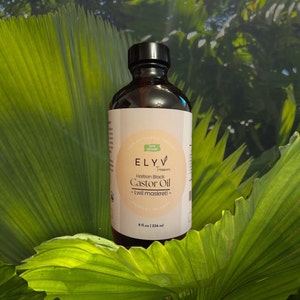 May include: A brown glass bottle with a black cap, labeled "ELYV Treasures Haitian Black Castor Oil - Lwil maskreti - Product of Haiti" and "8 fl. oz | 236 ml". The bottle is sitting on a bed of green leaves.